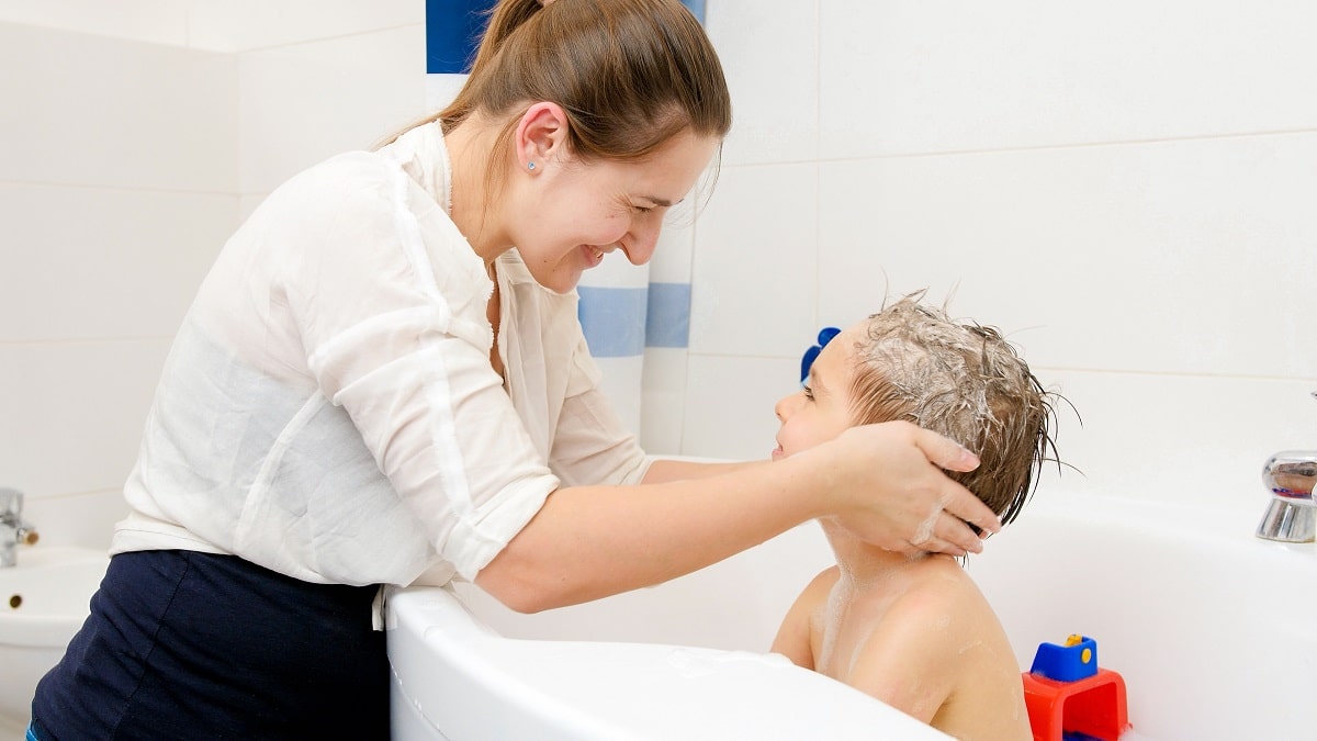 young smiling mother pouring shampoo on hand and washing hair of her little son sitting in bath. concept of child hygiene and health care at home. family having time together and playing at home