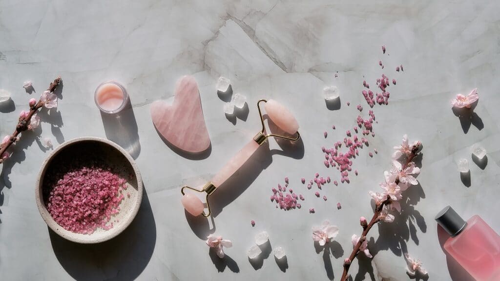 crystal rose quartz facial roller and gua sha stone for beauty facial massage therapy, flat lay on marble table with magnolia flowers. long shadows. essential oil bottle.
