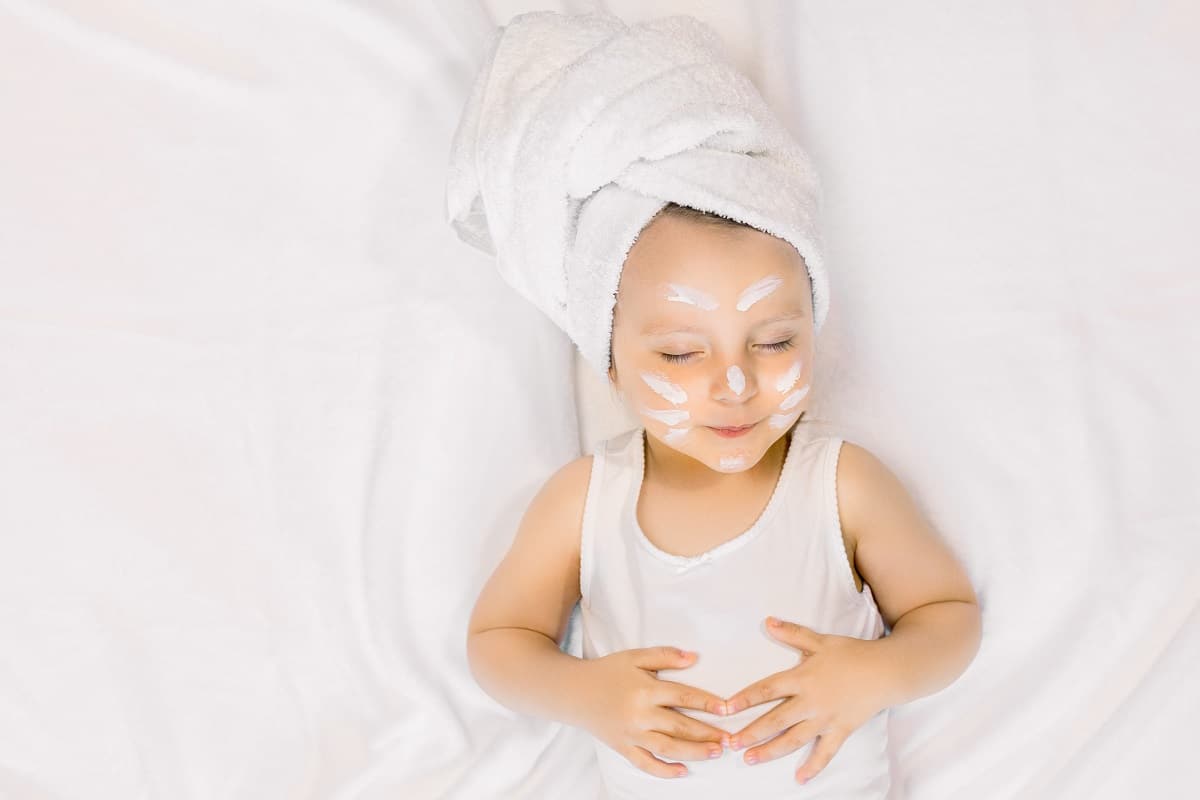photo of relaxed pretty little baby girl with cream on her face and bath towel on head, lying with closed eyes on white background after spa bath procedures. copy space