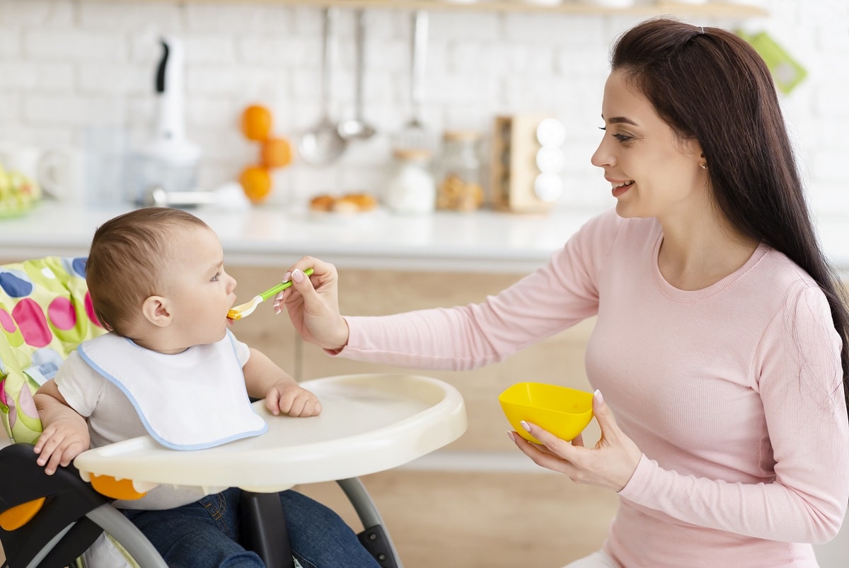mother feeding her baby, toddler sitting in high chair at kitchen