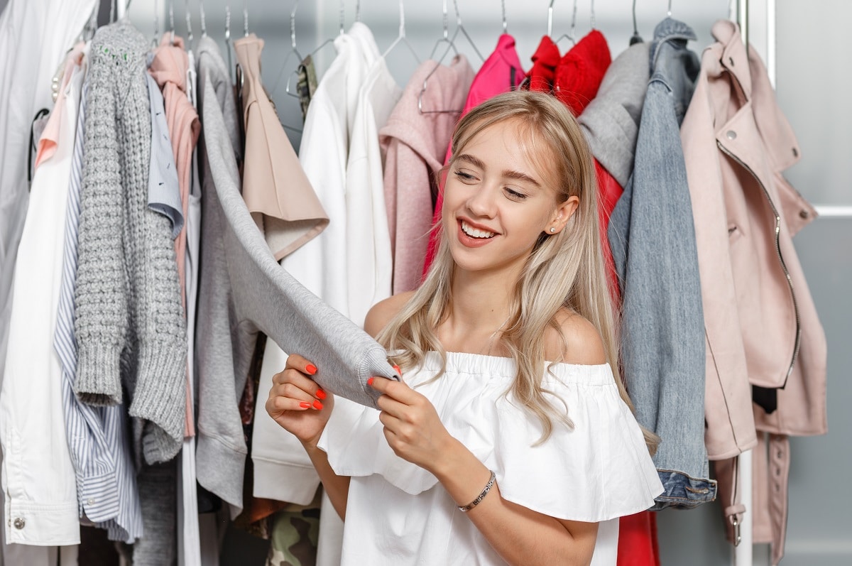 pretty girl blogger stands on the background of clothes hanging on a hanger in the wardrobe and touches the sleeve of the jacket