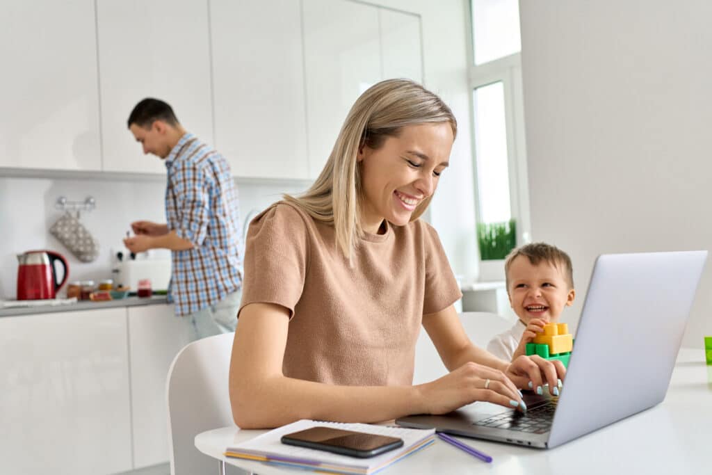 happy mom working at home using laptop while funny kid son playing.