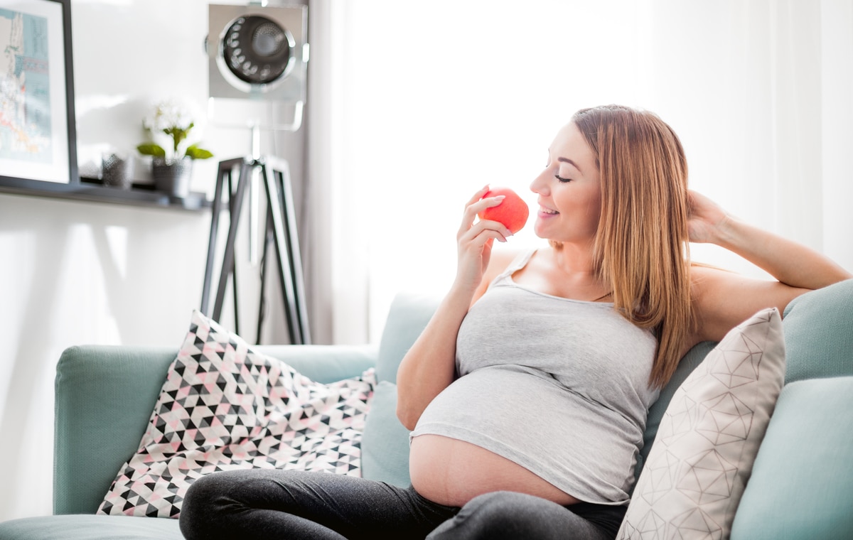 nutrition and diet during pregnancy, pregnant woman eating fruits sitting on sofa at home