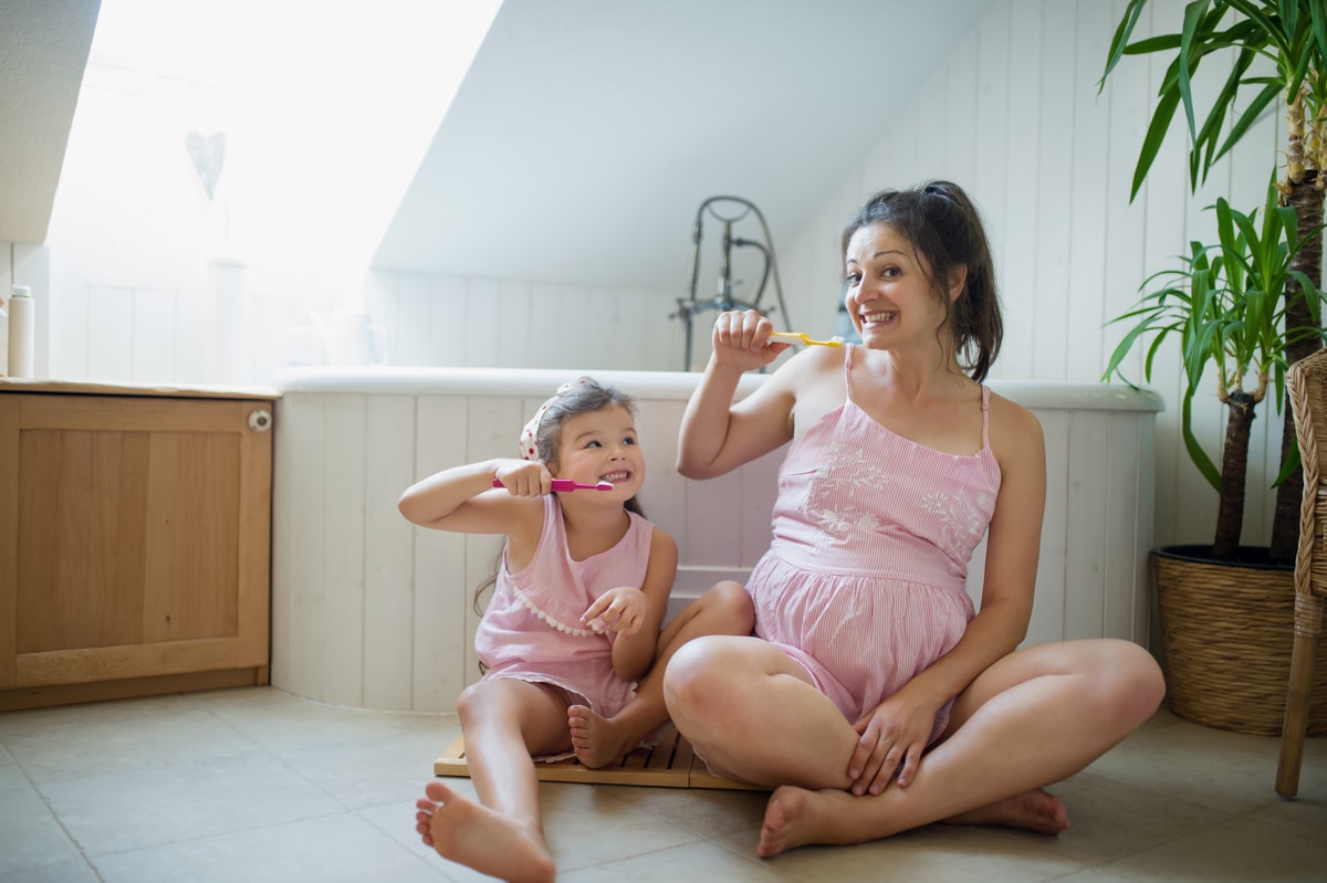 portrait of pregnant woman with small daughter indoors in bathroom at home, brushing teeth.