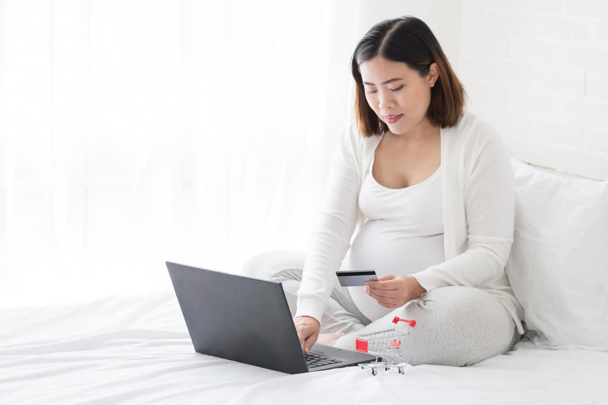 southeast asian pregnant women buy baby products from internet by black laptop and holding credit card in the white bedroom at home, pregnancy online shopping concept