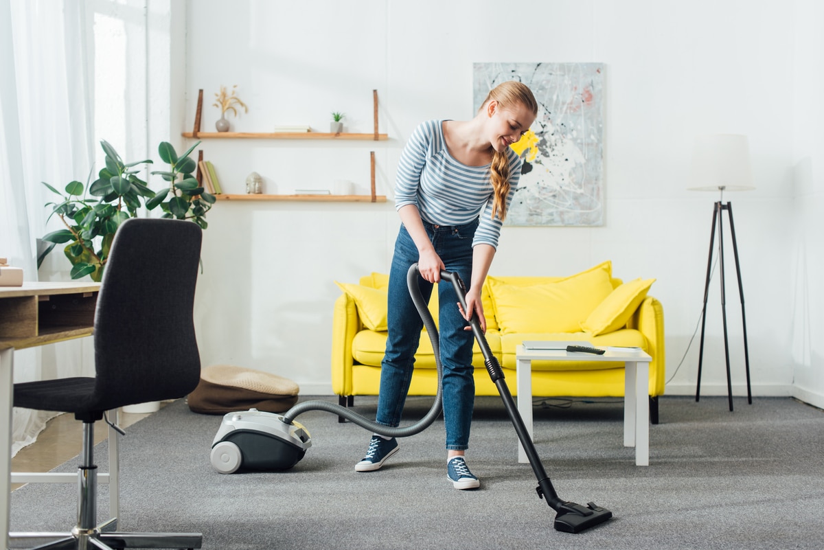 smiling woman cleaning carpet with vacuum cleaner in living room