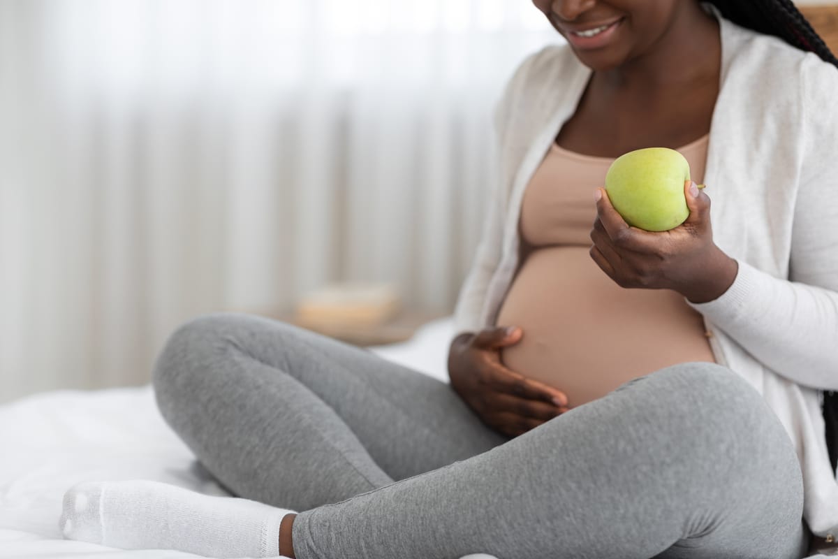 black pregnant woman eating green apple while sitting on bed at home