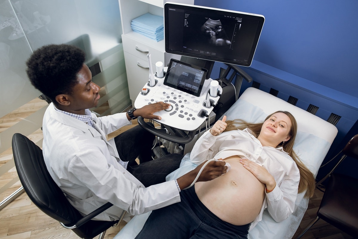 pregnancy and ultrasound concept. top angle view of young caucasian pregnant woman lying on the couch and showing thumb up, while high skilled male african american doctor making ultasound scan