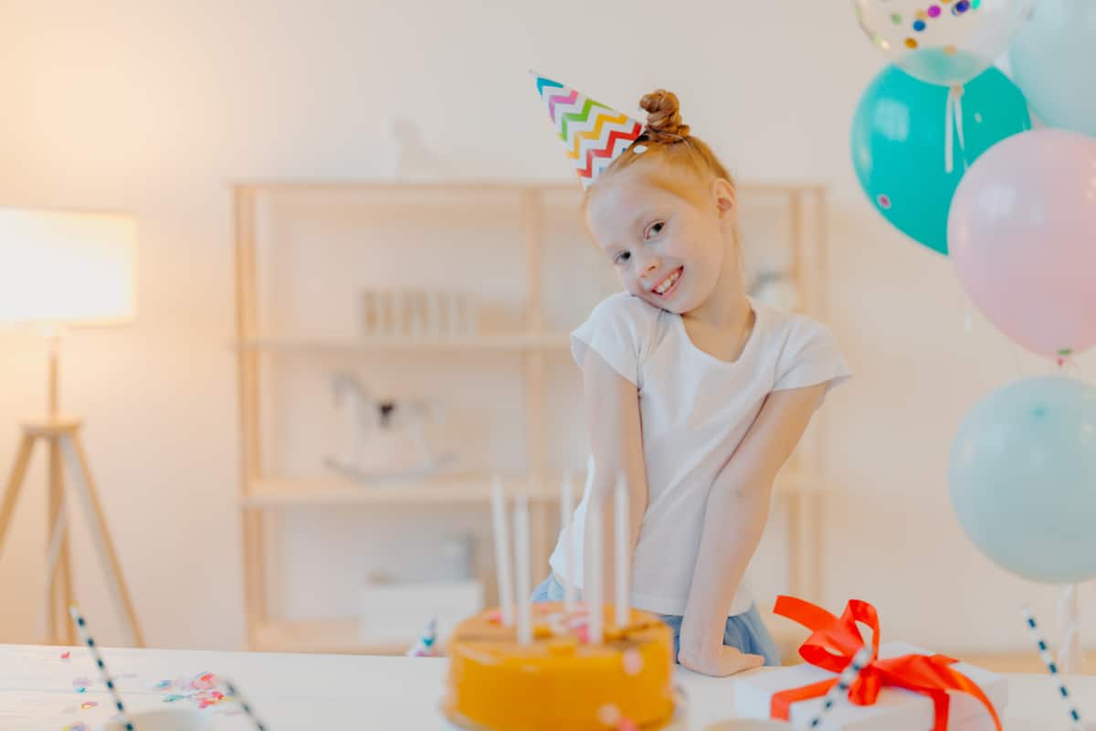 little ginger girl wears party hat and white tshirt, stands near festive table with cake, blows candles and makes wish during her birthday, poses in white room with inflated balloons, smiles joyfully