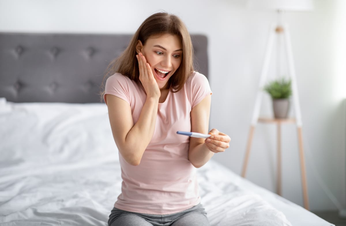 excited young woman checking her recent pregnancy test, feeling happy over future maternity on bed at home
