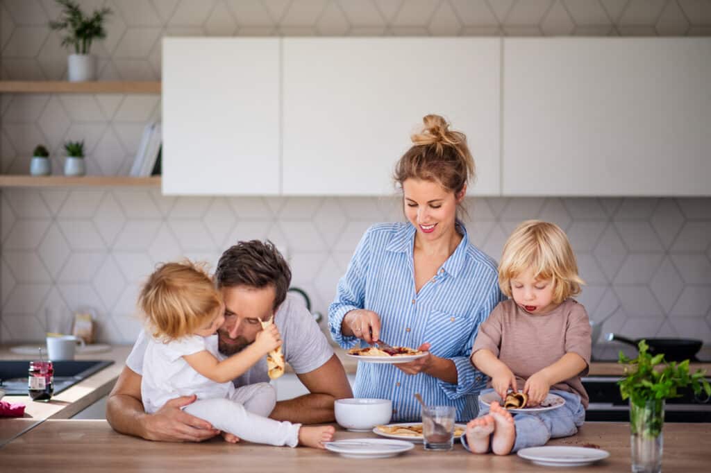 young family with two small children indoors in kitchen, eating pancakes.