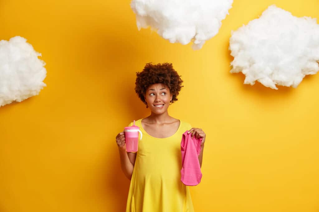 pensive pregnant young afro american woman dreams about child and future life holds feeding bottle and baby clothes bites lips looks thoughtfully away poses against yellow background with clouds