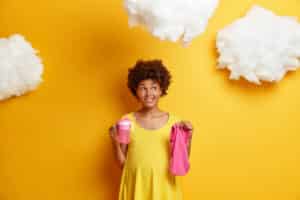pensive pregnant young afro american woman dreams about child and future life holds feeding bottle and baby clothes bites lips looks thoughtfully away poses against yellow background with clouds