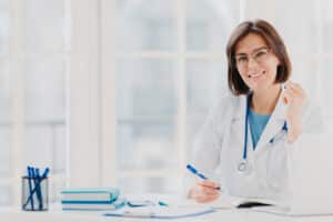 photo of woman intern works in hospital, writes in medical certificate, prescribes treatment for patient, makes recipe, dressed in white coat with phonendoscope, leaves notes of medical examination