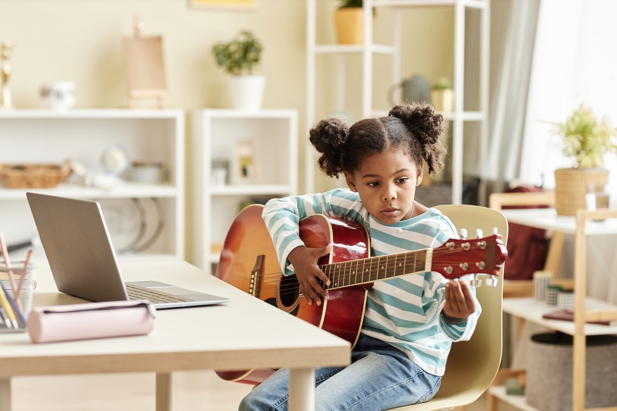 black little girl playing guitar at home and watching online music lesson