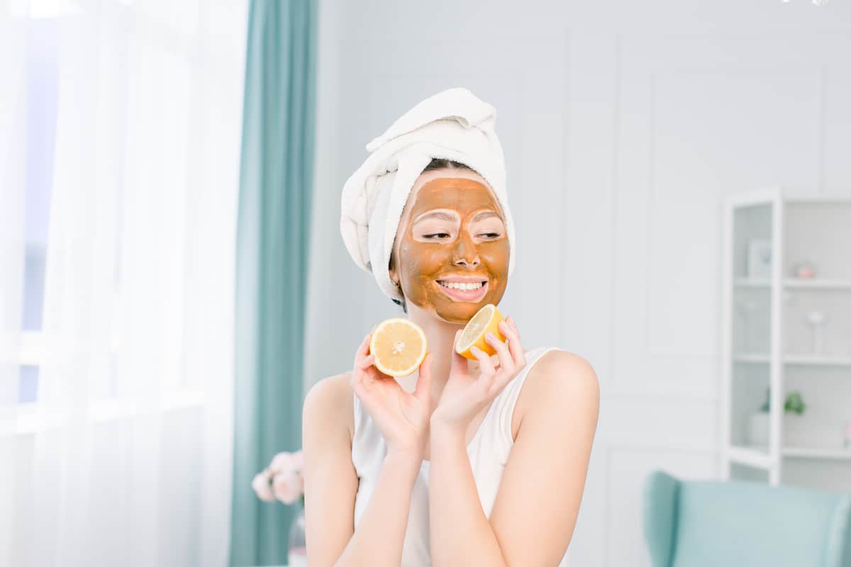 beauty procedures skin care concept. young woman with brown facial mud clay mask on her face in bathroom, with white towel on head, holding lemon halfs