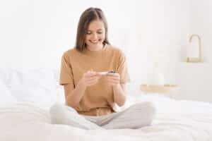 young woman sitting on bed, looking at pregnancy test showing positive result, smiling happily