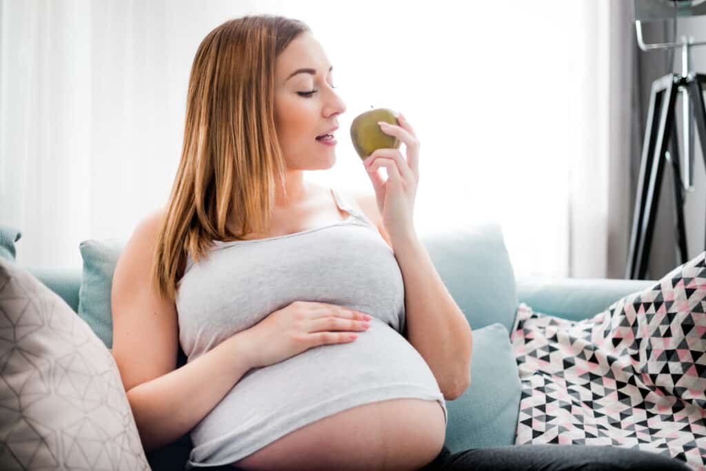 nutrition and diet during pregnancy, pregnant woman eating fruits sitting on sofa at home