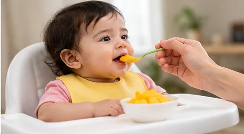 Baby eating mango from a spoon while seated in a high chair